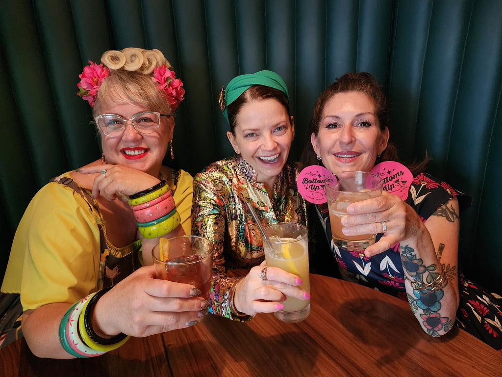 Three women sitting together, smiling and holding drinks against a dark curtain background.