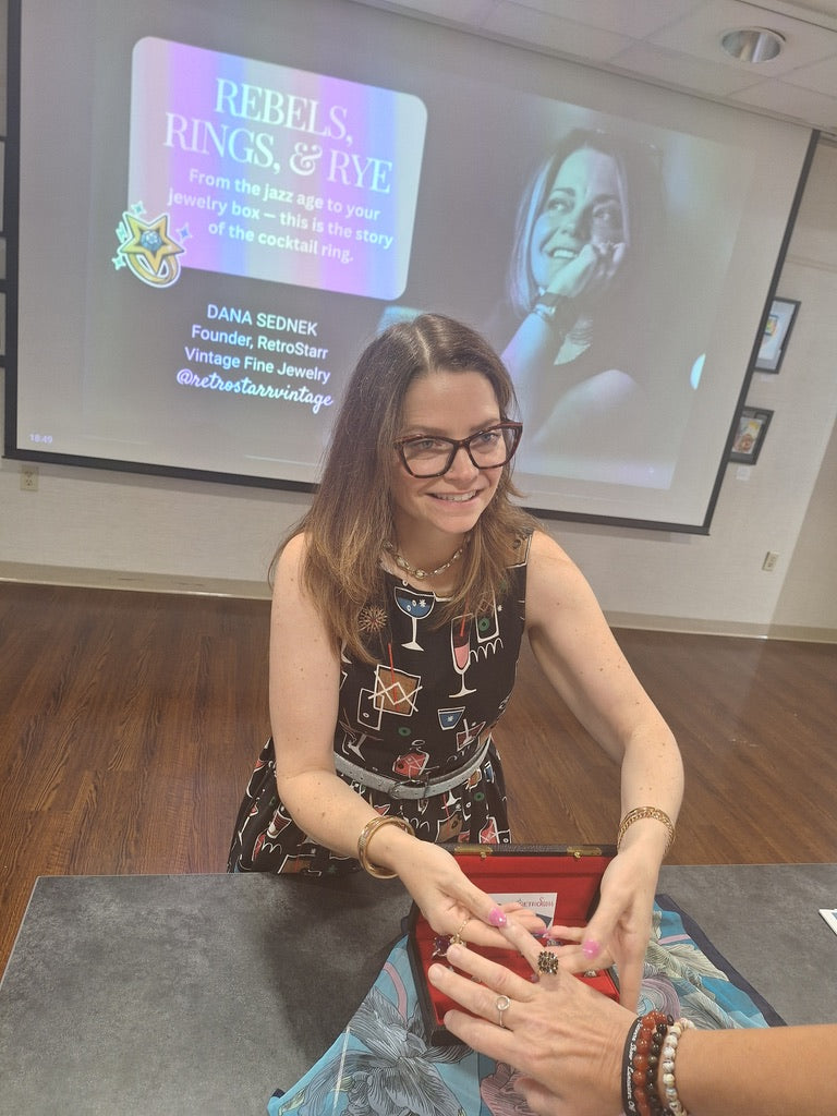 Woman signing an item at a table with a screen displaying 'Rebels, Rings, & Rye' in the background.