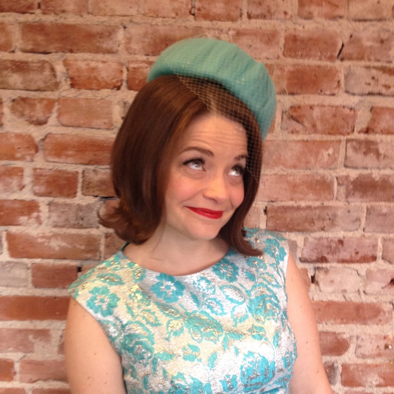 Woman wearing a teal dress and beret against a brick wall